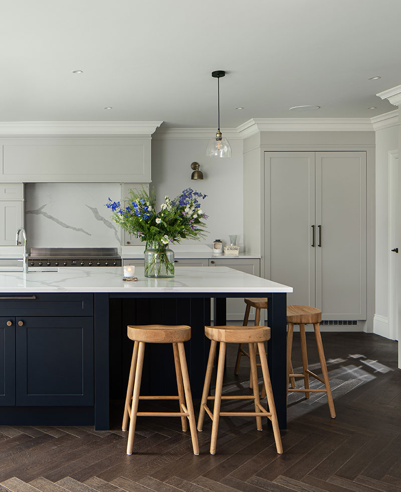 Bright, airy kitchen with dark blue island, white marble countertops, and light gray cabinetry. A bouquet of wildflowers sits on the island, and three wooden stools are tucked underneath. Hardwood floors with a herringbone pattern complete the space.