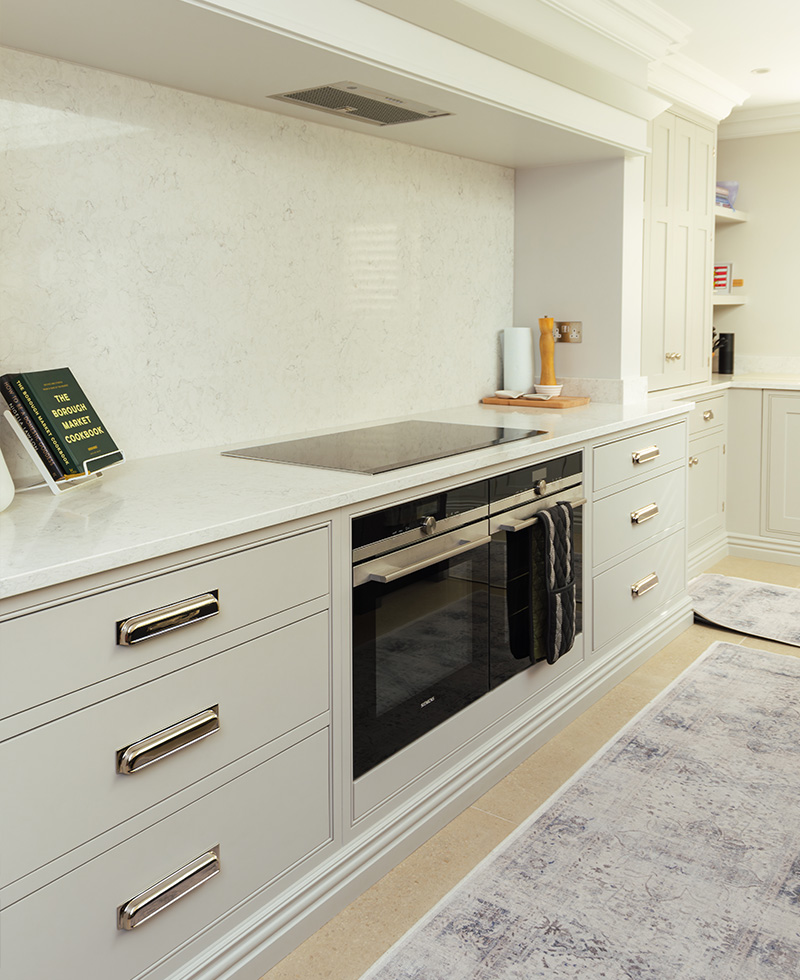 A modern kitchen with white marble countertops, light grey cabinets, and stainless steel appliances. A cookbook rests on the counter, and a patterned rug covers the floor.